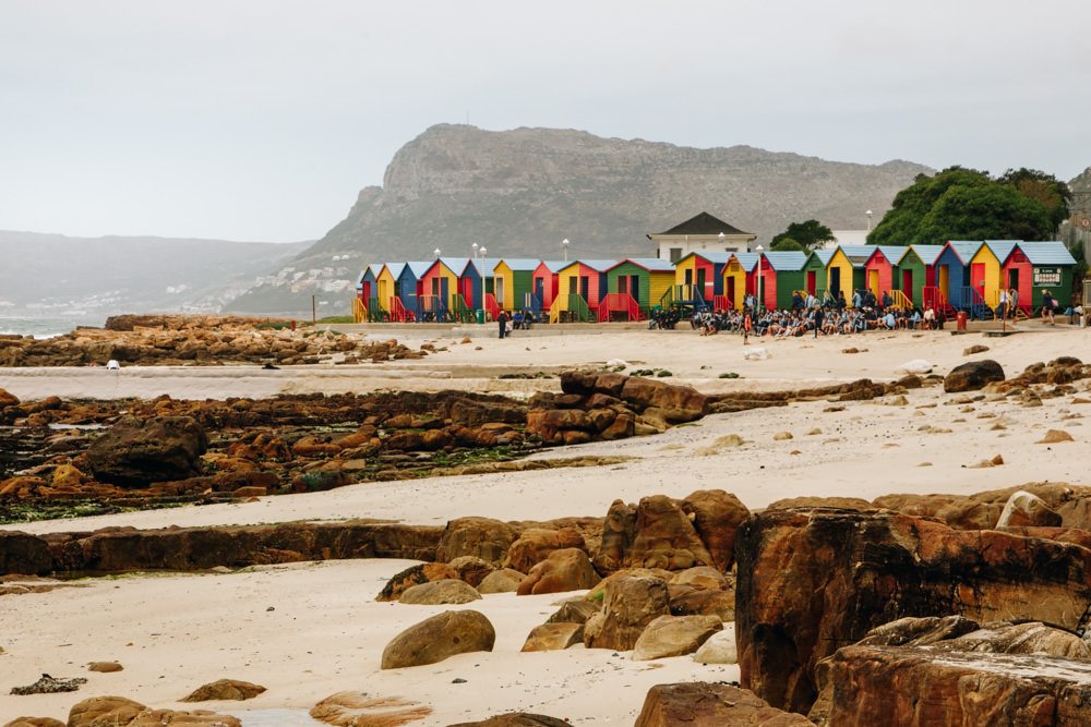 Colourful beach huts in a row with a sand and rocky beach in front
