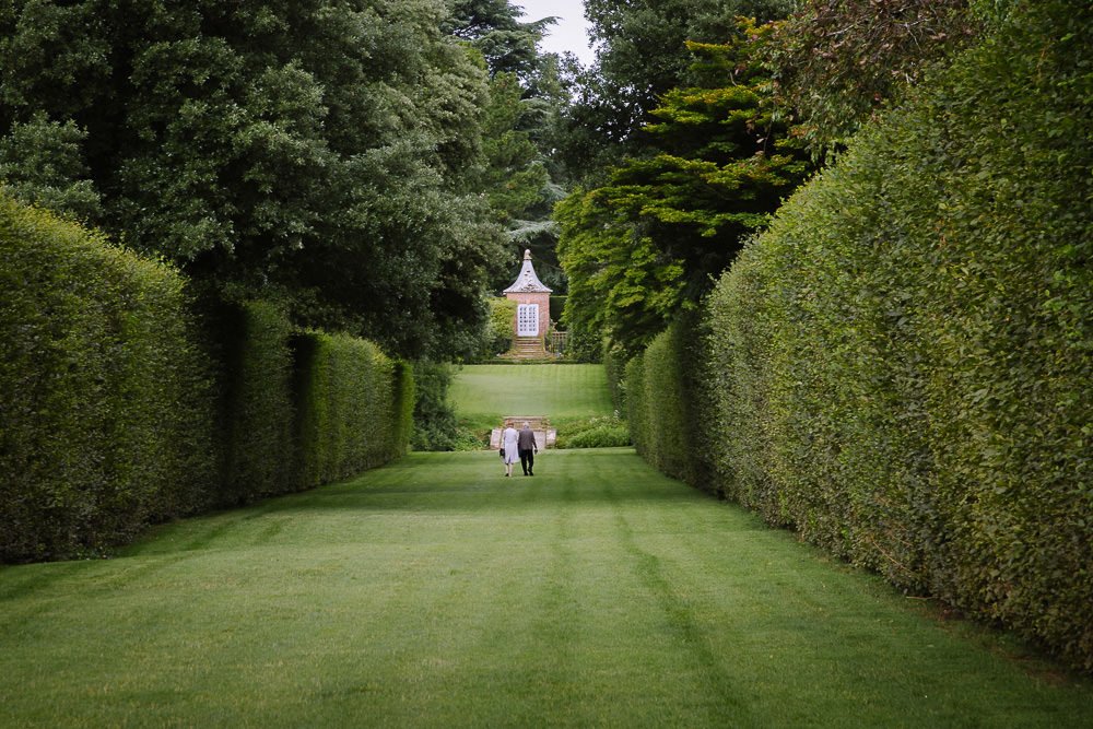 Couple walk over manicured lawn between high hedges
