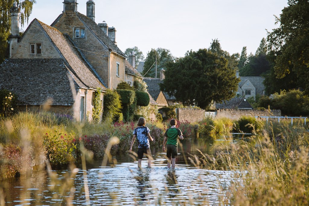 Two people walk through a river beside stone cottages on a late summer afternoon